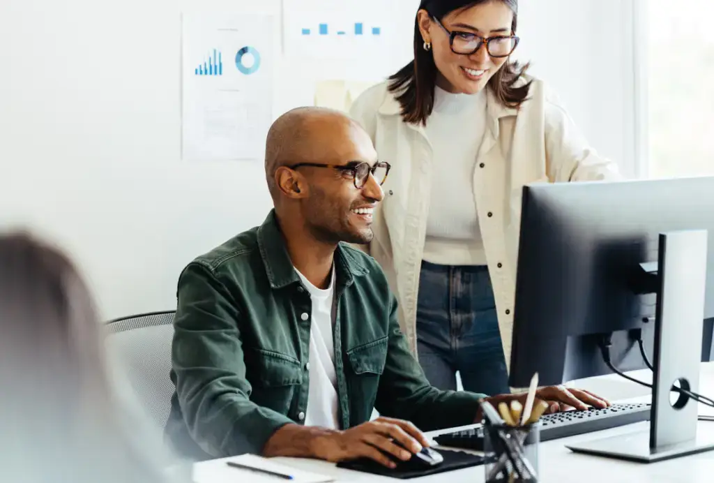 A man and woman collaborate at a desk, with the man smiling as the woman points to the computer screen in a calm office setting.